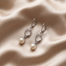Silver Flower Engraved Crystal & Round Faux Pearl Drop Earrings displayed on a draped beige satin fabric background.