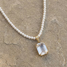 A pearl necklace with a gold-toned pendant featuring a clear octagon shaped crystal, displayed on a textured gray stone surface.