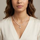 A close-up of a woman wearing a delicate white pearl necklace with a small gold pendant featuring a clear faceted gemstone, paired with a cream-colored satin top.