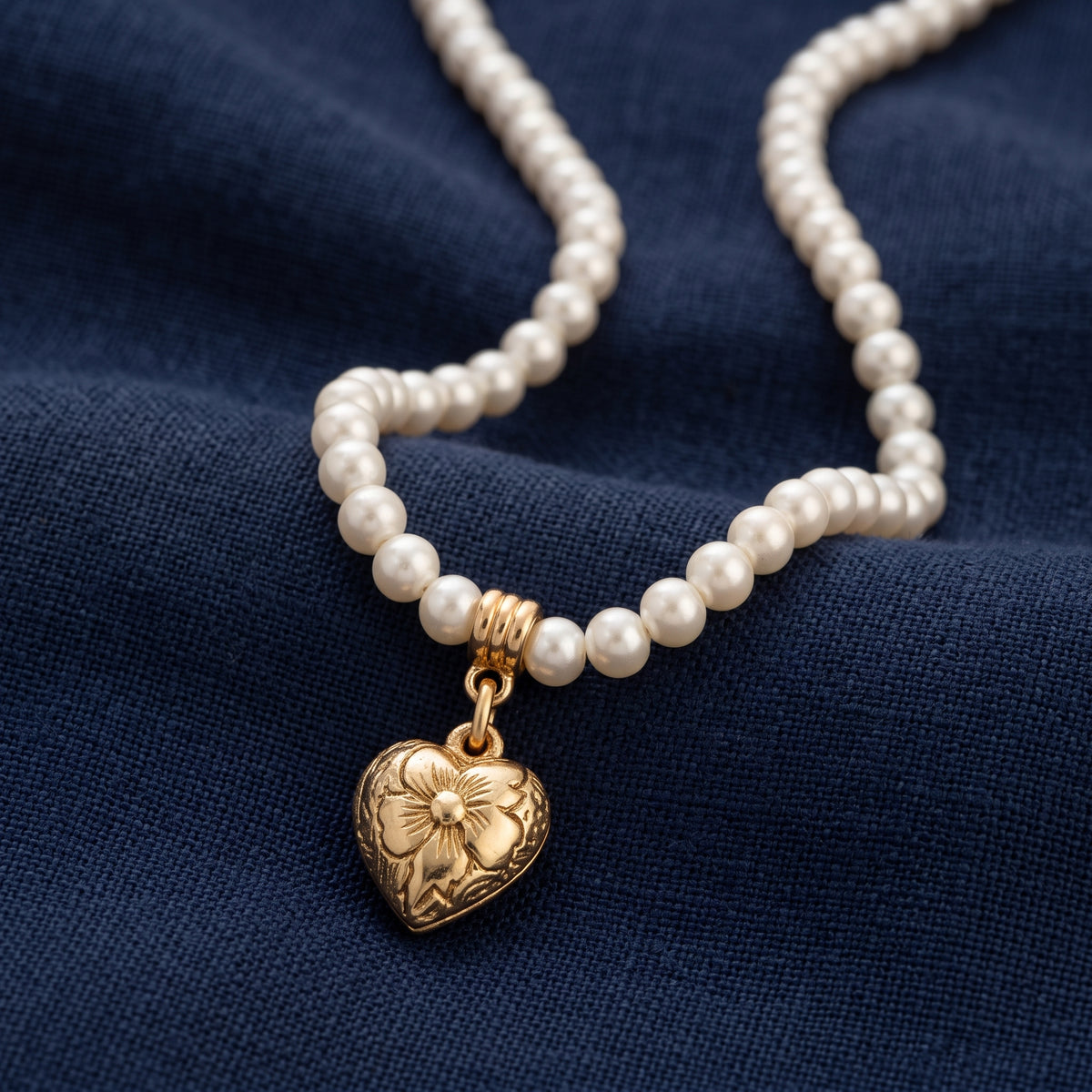 Gold tone etched heart pendant pearl strand necklace displayed on a textured navy blue fabric background.