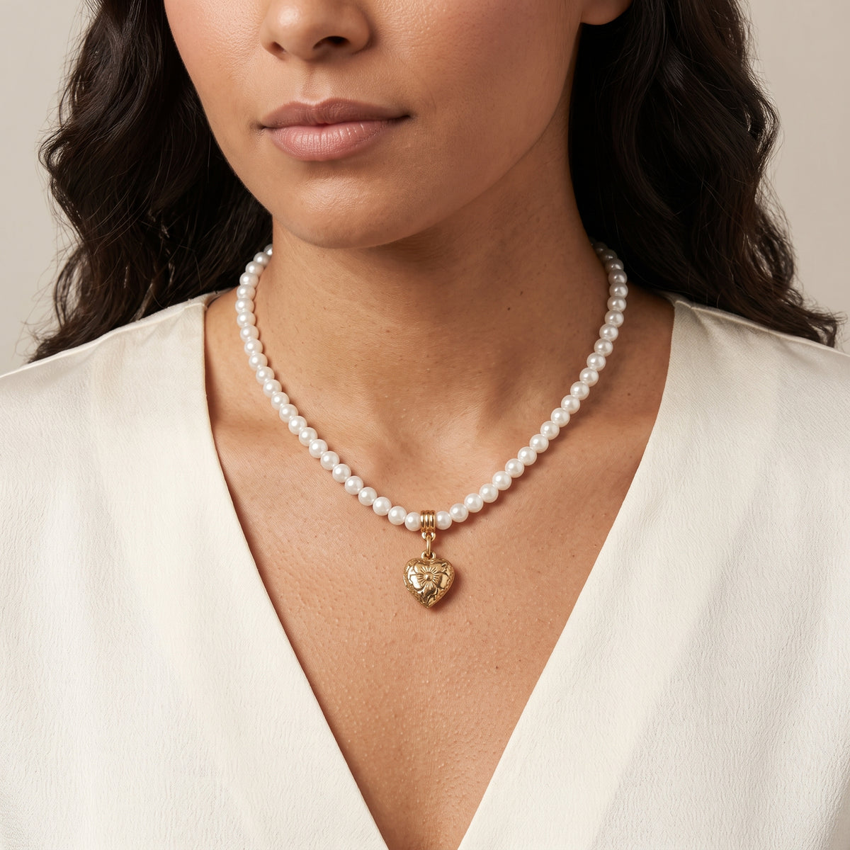 A close-up of a model wearing a Floral Gold Tone Etched Heart Pendant & Round Faux Pearl Beaded Strand Necklace, styled with a white V-neck top against a soft beige background.