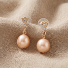 Golden Pink Champagne Faux Pearl & Crystal Drop Stud Earrings displayed on a textured beige fabric background.