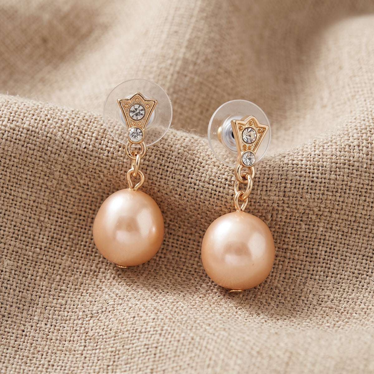 Golden Pink Champagne Faux Pearl & Crystal Drop Stud Earrings displayed on a textured beige fabric background.