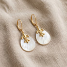 Oval Mother of Pearl & Gold Fleur De Lis Drop Earrings resting on beige linen cloth