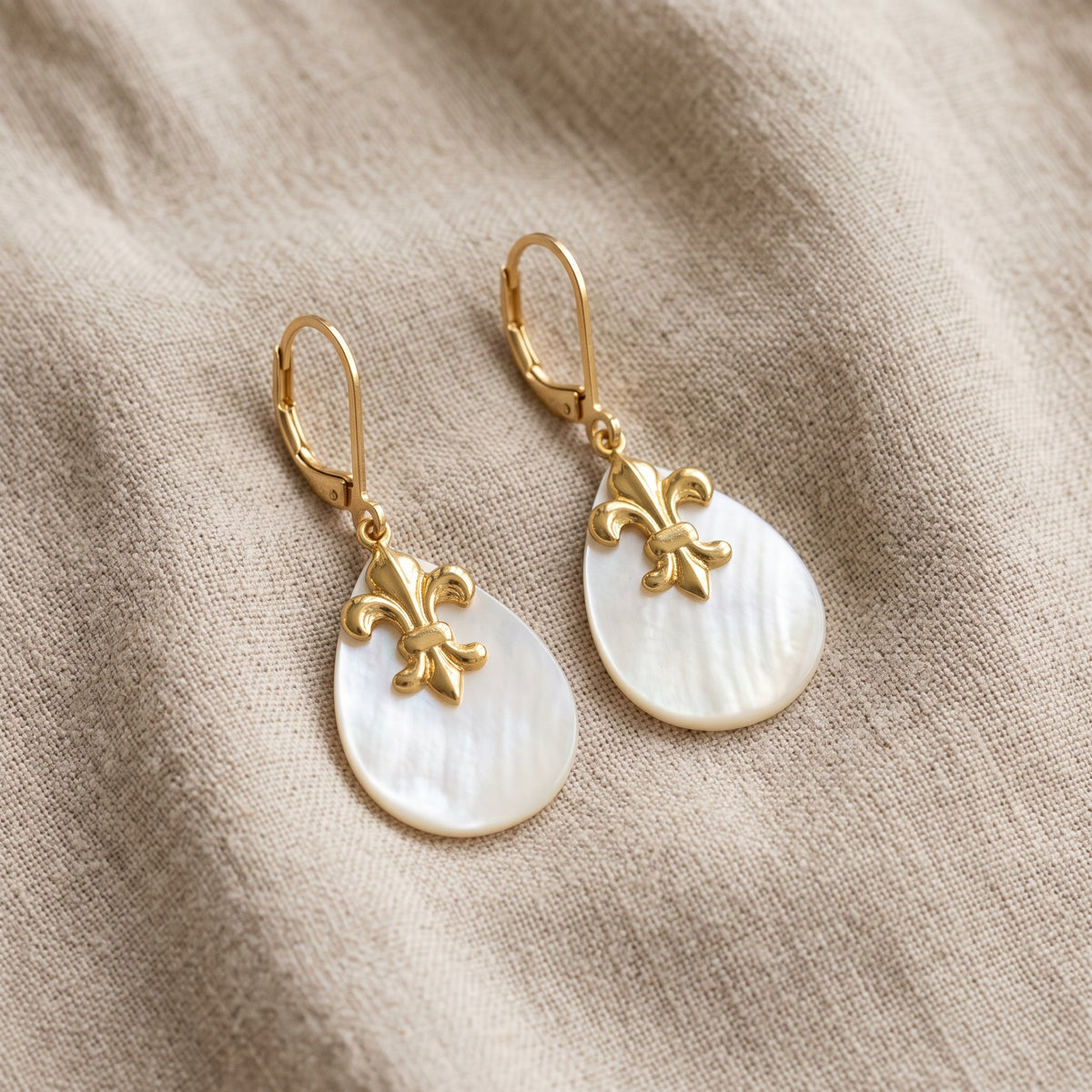 Oval Mother of Pearl & Gold Fleur De Lis Drop Earrings resting on beige linen cloth