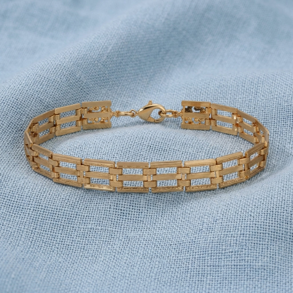 A gold tone metal link bracelet with a geometric, rectangular openwork design, resting on a textured light blue fabric background.