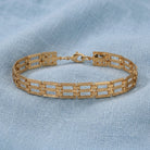 A gold tone metal link bracelet with a geometric, rectangular openwork design, resting on a textured light blue fabric background.