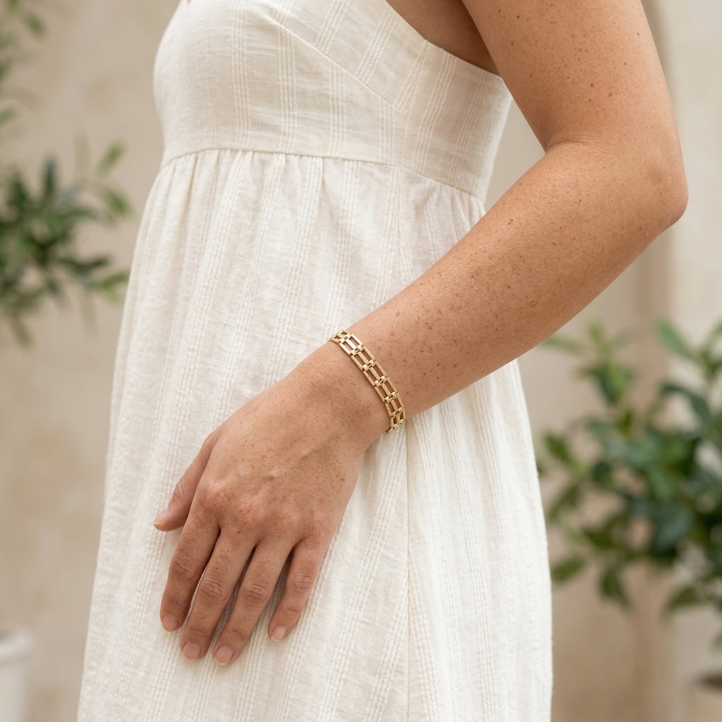 A close-up of a person wearing a textured cream-colored dress and a gold chain link bracelet, with a soft-focus background of green plants.