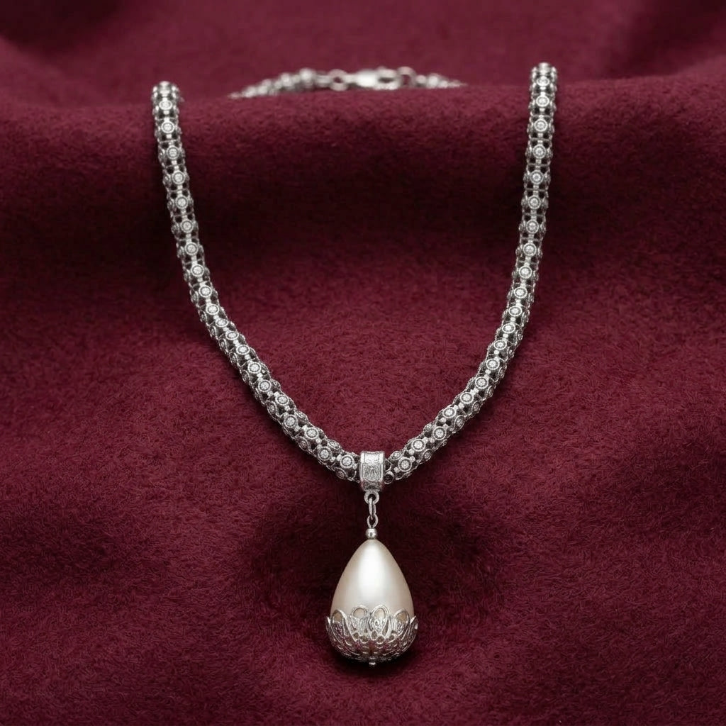A silver-toned necklace with a textured pavé chain and a teardrop-shaped pearl pendant featuring a cream-colored pearl-like center, displayed on a deep burgundy velvet fabric background.
