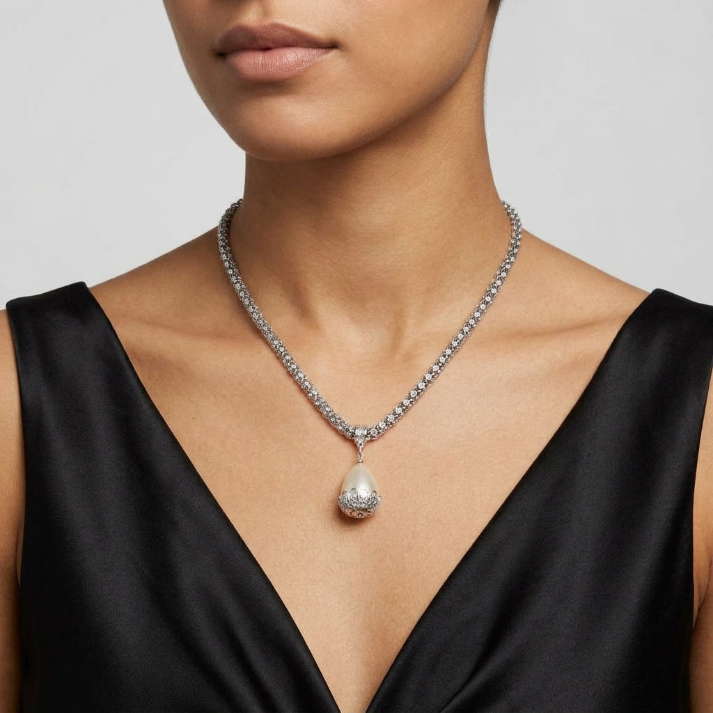 A close-up of a model wearing a silver-toned crystal necklace with a teardrop-shaped pearl pendant, paired with a black satin V-neck top against a light gray background.
