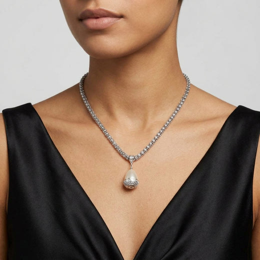 A close-up of a model wearing a silver-toned crystal necklace with a teardrop-shaped pearl pendant, paired with a black satin V-neck top against a light gray background.