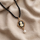 A black beaded necklace with a vintage-style oval pendant featuring an ivory-colored classical cameo portrait, surrounded by a gold-tone frame with small clear stones and a dangling faux pearl, displayed on draped beige linen fabric.