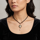 A close-up of a model wearing a black beaded necklace with a vintage-style cameo pendant in a gold-toned frame and a dangling faux pearl drop.