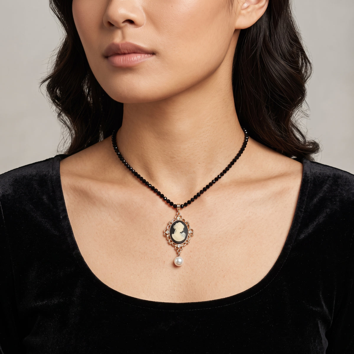 A close-up of a model wearing a black beaded necklace with a vintage-style cameo pendant in a gold-toned frame and a dangling faux pearl drop.
