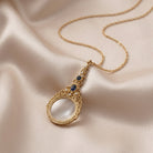 Gold necklace with a magnifying glass pendant accented with Montana Blue on a beige fabric background