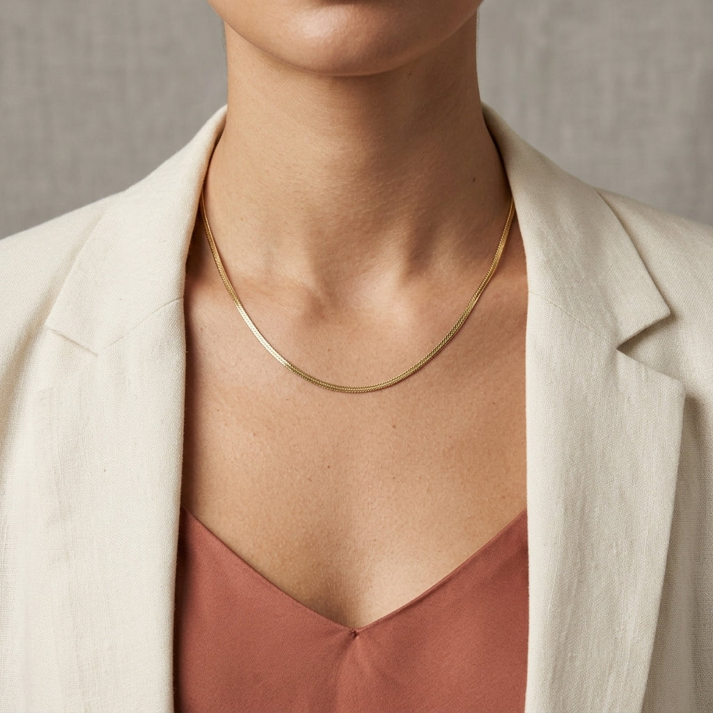 A close-up of a model wearing a delicate, thin gold snake chain necklace, styled with a cream-colored blazer and a dusty rose top.