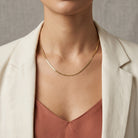 A close-up of a model wearing a delicate, thin gold snake chain necklace, styled with a cream-colored blazer and a dusty rose top.