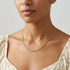 A close-up of a model wearing a delicate gold chain necklace with a small bead link design, paired with a cream-colored floral top against a light gray background.