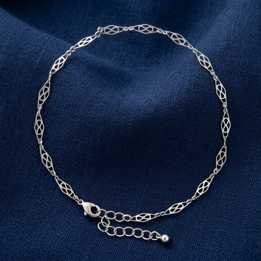 A silver chain anklet with an intricate openwork link pattern, displayed on a textured navy blue fabric background.