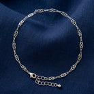 A silver chain anklet with an intricate openwork link pattern, displayed on a textured navy blue fabric background.