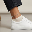 Woman wearing a silver tone twisted chain anklet with white sneakers and black denim jeans