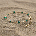 A delicate gold chain anklet with small faceted green crystal beads, displayed on a textured sandy surface.