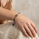 A close-up of a person's hand resting on a textured beige surface, wearing a bohemian-style Black Diamond Fireball Crystal Brown Leather Cord Linen Wrapped Bracelet
