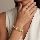 A close-up of a woman wearing a gold and pearl bracelet on her wrist, with her hand resting near her neck. She is wearing a cream-colored sleeveless top against a soft beige background.