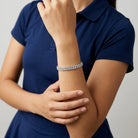 A close-up of a model wearing a slim rhinestone bracelet on her wrist, paired with a navy blue polo shirt against a neutral gray background.