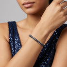 A close-up of a model wearing a delicate silver-toned and blue crystal rhinestone bracelet, paired with a sparkling navy blue sequined top against a neutral gray background.