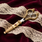 Manor House Pink And Multicolor Flower Decal Cherub Scissors resting on a deep burgundy velvet fabric with cream lace.