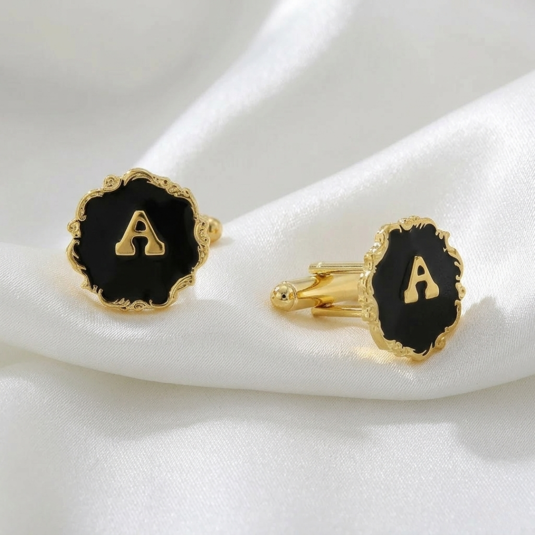 Gold cufflinks with black enameled discs featuring the letter 'A' on a white fabric background.