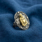A silver-toned ring featuring a raised gold-toned Buddha figure, displayed on a textured blue fabric background.