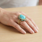 Hand wearing a turquoise ring with gold chain on a wooden surface