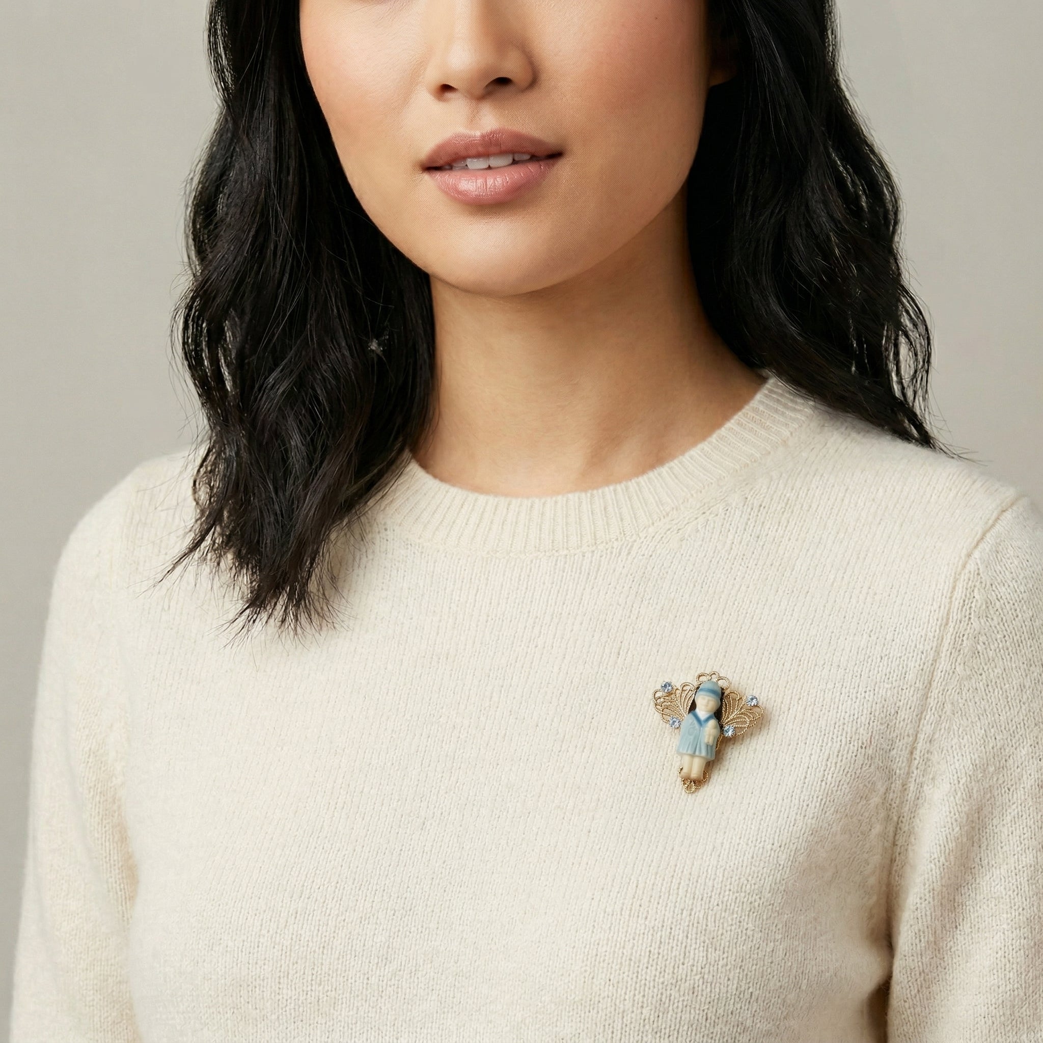 Woman wearing a beige sweater with a blue crystal Winged Filigree Porcelain Child With Doll Pin