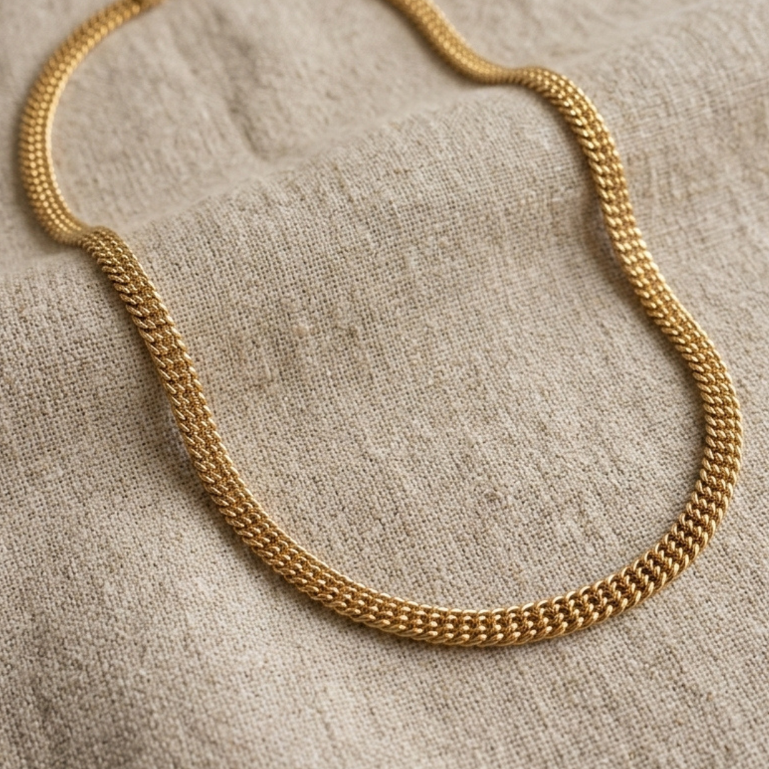 A gold-toned chain necklace with a textured, woven-like link pattern, draped on a neutral beige linen fabric background.