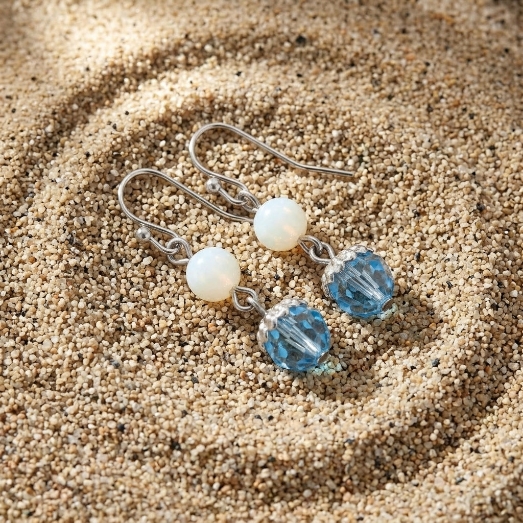 A pair of dangle earrings resting on sand, featuring silver-toned fish hook wires, smooth white round beads, and faceted light blue oval drops.