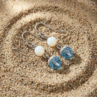 A pair of dangle earrings resting on sand, featuring silver-toned fish hook wires, smooth white round beads, and faceted light blue oval drops.