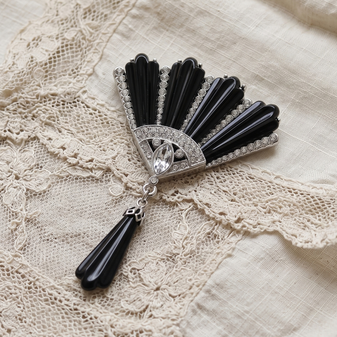 A vintage-style Art Deco brooch featuring black onyx fan-shaped elements, a silver-toned central frame with clear crystals, and a dangling black onyx tassel, displayed on a cream lace fabric background.