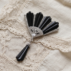 A vintage-style Art Deco brooch featuring black onyx fan-shaped elements, a silver-toned central frame with clear crystals, and a dangling black onyx tassel, displayed on a cream lace fabric background.