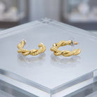A pair of gold twisted rope hoop earrings displayed on a clear acrylic jewelry stand against a softly blurred gray background.