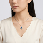 Woman wearing a necklace with a blue "B" initialed pendant on a plain background