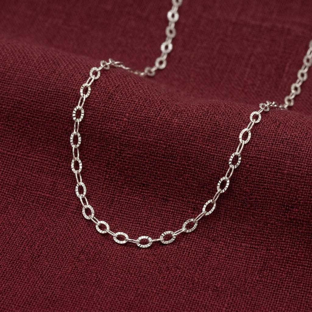 A silver-toned chain necklace with repeating oval links, draped on a textured dark red fabric background.