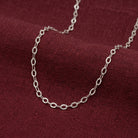 A silver-toned chain necklace with repeating oval links, draped on a textured dark red fabric background.
