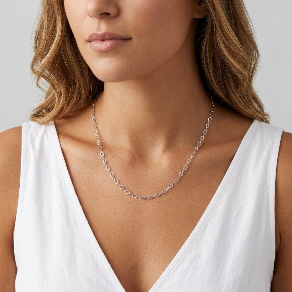 A close-up of a woman wearing a delicate silver chain necklace with small oval links, styled with a white sleeveless top against a light gray background.