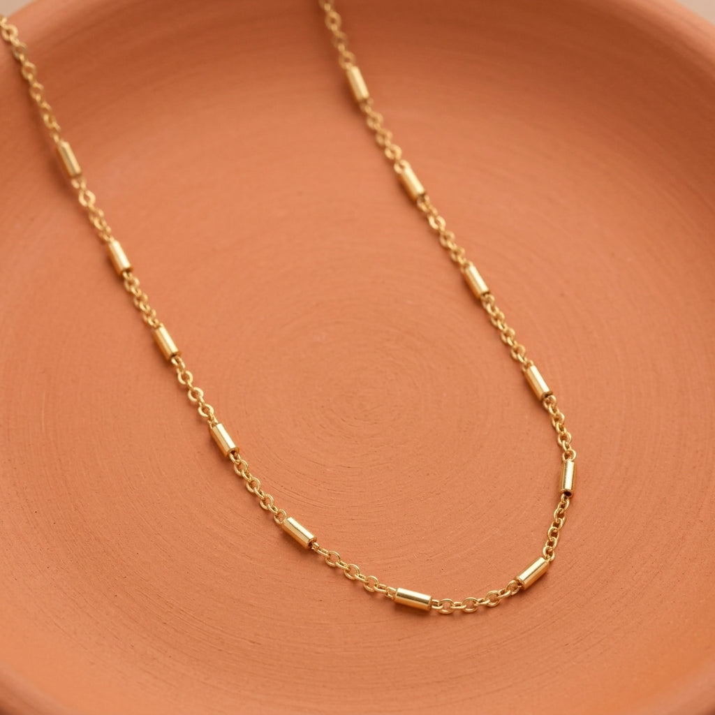 A delicate gold chain necklace with cylindrical links, draped elegantly on a textured terracotta-colored surface.