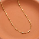 A delicate gold chain necklace with cylindrical links, draped elegantly on a textured terracotta-colored surface.