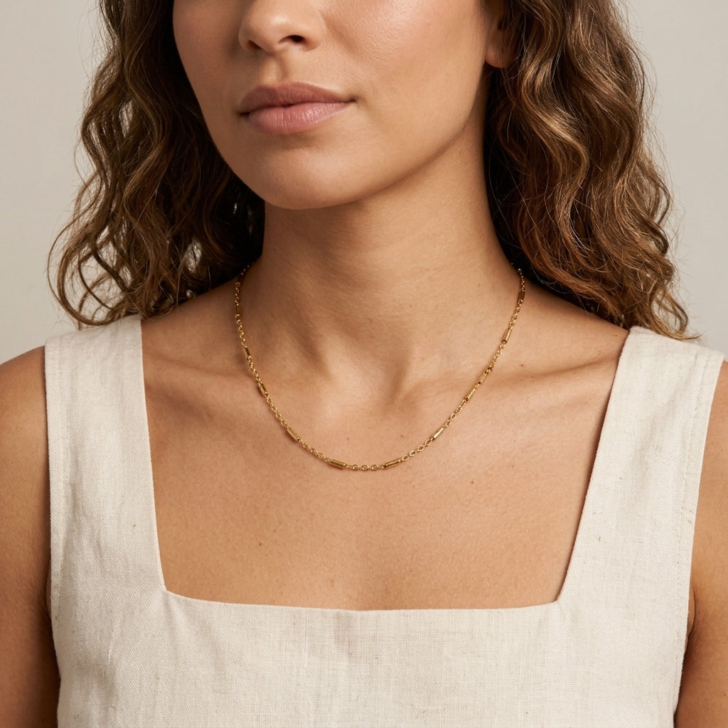 A close-up of a woman wearing a delicate gold chain necklace with a cream linen top against a neutral beige background.