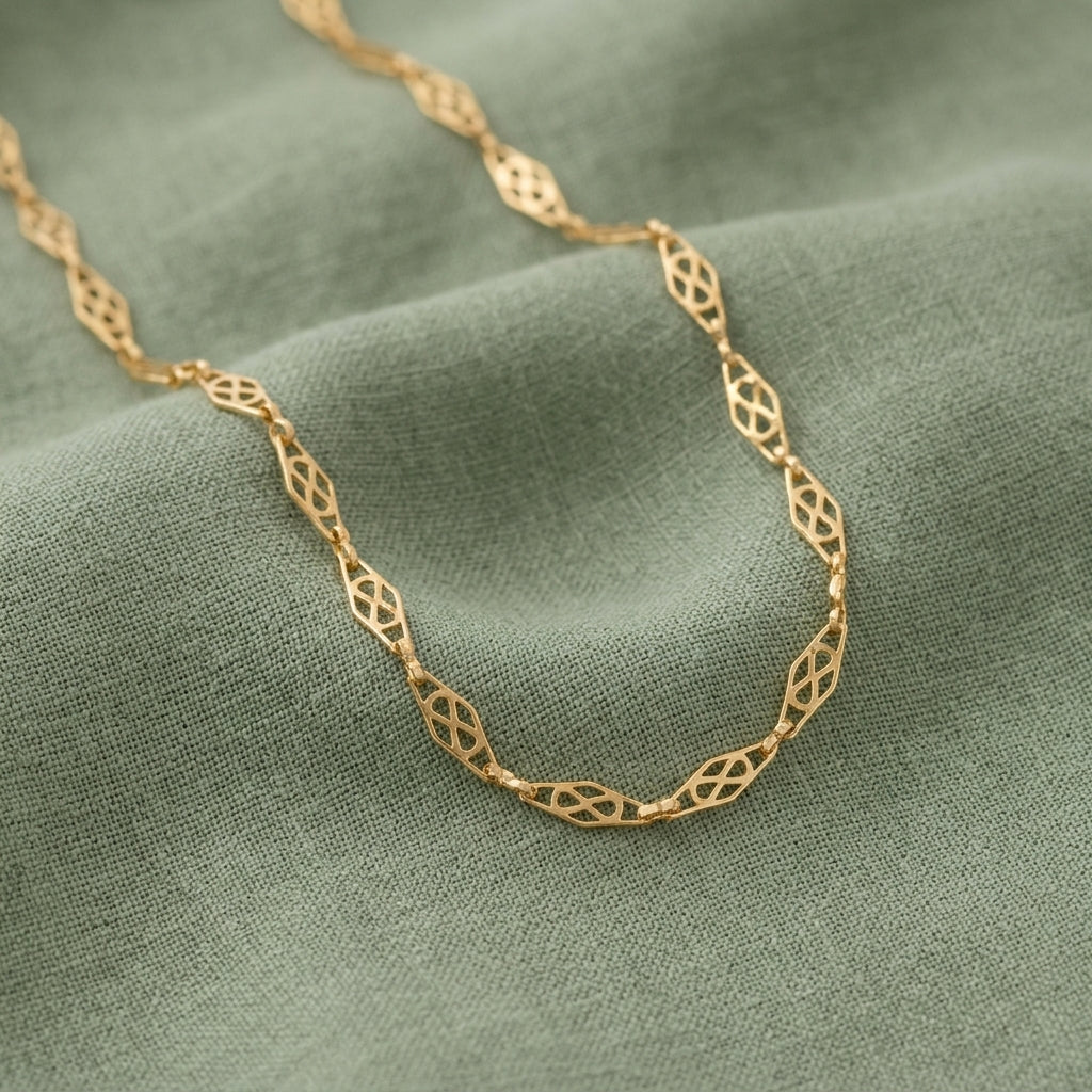 A gold chain necklace with an openwork geometric link pattern, draped elegantly on a textured sage green fabric background.
