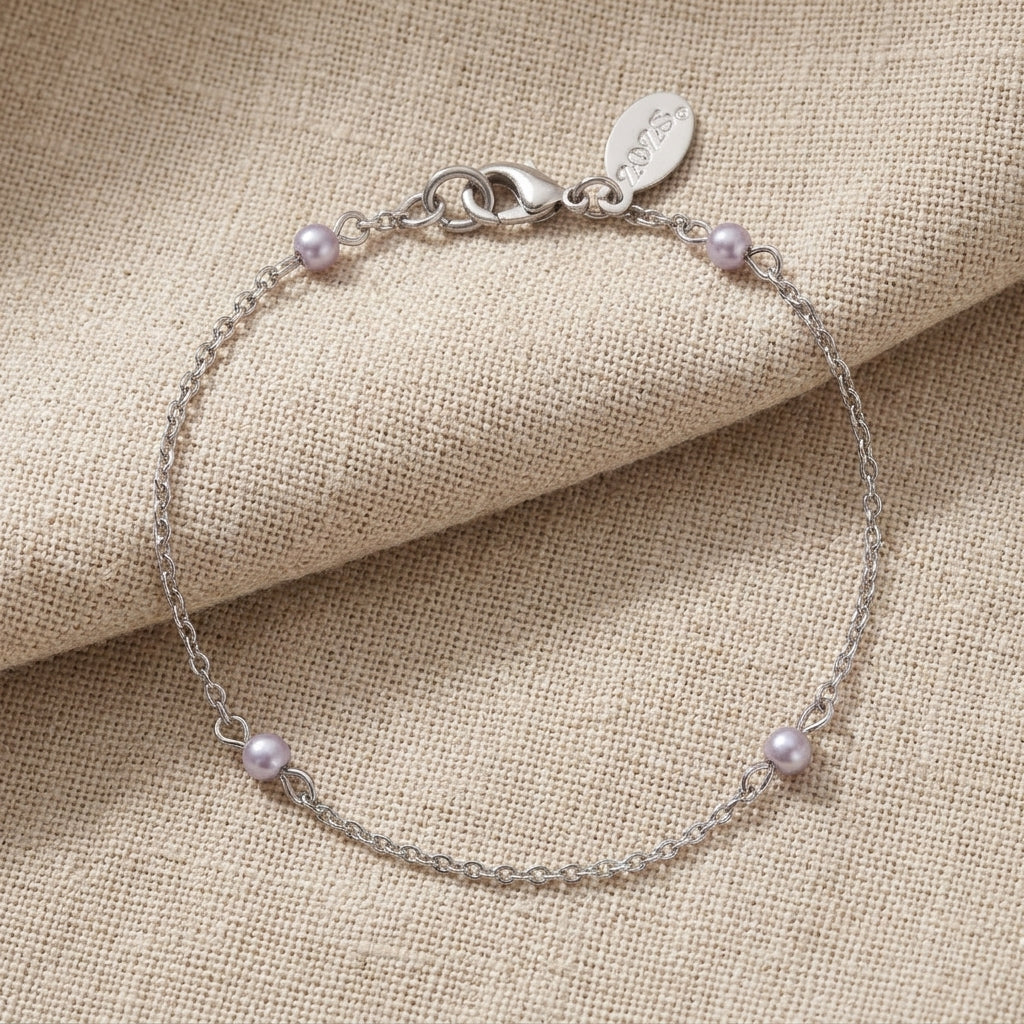 A delicate silver chain bracelet with four small light gray pearls spaced along the chain, displayed on a textured beige fabric background. The clasp is a lobster clasp with an engraved tag.